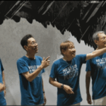 青いTシャツを着た4人のバンドメンバーが横を指さして笑顔で並んでいる様子